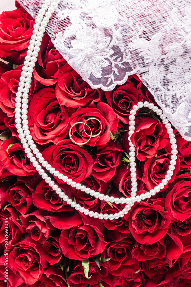 Obraz premium Wedding composition for the design of a postcard, cover, invitation. Pearl beads, two gold rings against a background of gorgeous red roses. Top view.