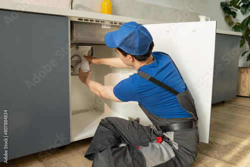 A skilled plumber in a blue uniform and cap is working diligently to repair the pipes located under the sink in a well-lit, contemporary kitchen setting, equipped with various tools for the job.