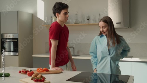 A young couple is engaged in a heated argument in a well-lit, contemporary kitchen setting. Tomato slices and a cucumber lay on a cutting board, suggesting dinner preparation was underway before the photo