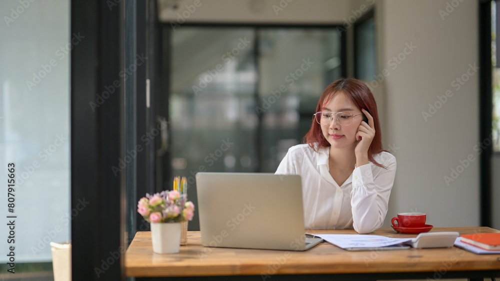 A woman is sitting at a desk with a laptop and a cup of coffee. She is wearing glasses and she is focused on her work A woman is sitting at a desk with a laptop and a cup of coffee. She is wearing glasses and she is focused on her work