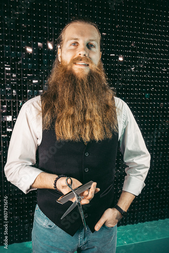 Bearded barber portrait with scissors .Man young barber hairdresser professional with long beard and hair in vest with scissors in hands portrait, cutting with scissors on black background. profession