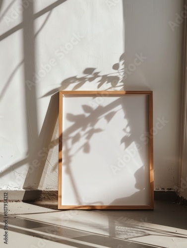  A thin, light wooden A4 frame is leaning against the wall on the floor, with white walls and sun rays shining in through the window above, making shadows across the room in a mini