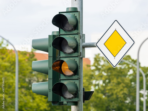 Traffic light blinking yellow and priority road sign next to it. The light signal is broken of switched off. The car drivers must drive according to the analog symbols.