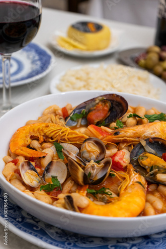 A dish of seafood feijoada with rice, garnished with shrimp and clams, sits on a restaurant table. Alongside are Alentejo cheese and a glass of red wine, evoking a warm Portuguese dining experience. photo