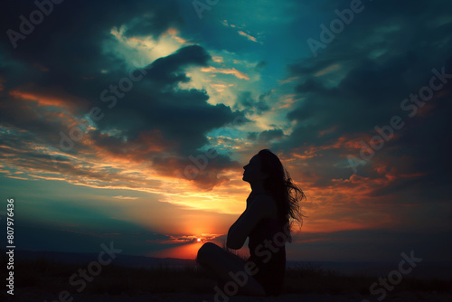 A woman in silhouette meditates while seated, with a vibrant sunset sky in the background, depicting calmness and inner peace