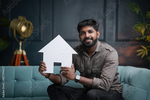 Smiling young Indian guy sitting on sofa and holding house, House loan and Reals state business concept image © sarath