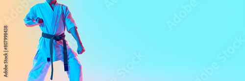 Cropped image of boy, karate athlete in uniform, green belt standing against gradient orange blue background in neon light. Concept of sport, martial arts, combat sport, healthy and active lifestyle photo