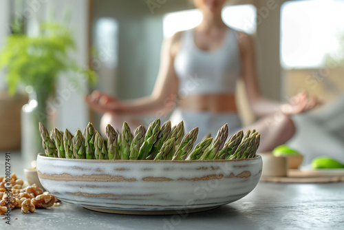 A young woman in a white sports bra and leggings meditates and doing yoga in a sunny room, with a bowl of asparagus and fruits nearby. Healthy living and mindfulness for wellness content. photo