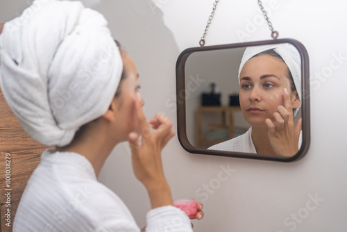 Home skincare: A woman, her reflection in the mirror, diligently applies face moisturizer, elevating her routine. 