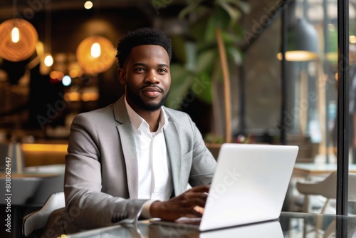 A young African-American professional is sitting in a cafe working on his laptop. He is wearing a suit and has a confident smile on his face.