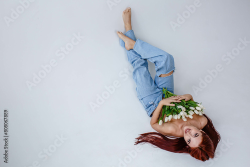 Red-haired girl in jeans with white tulips posing while lying on a white background