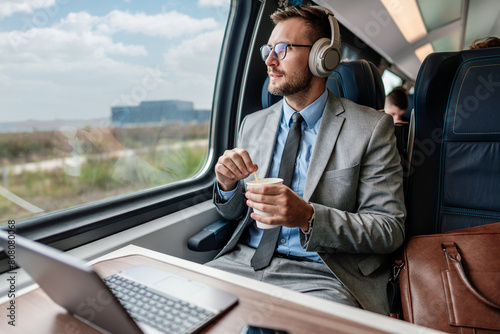 Handsome businessman is having a good time while traveling by high-speed train. He is using laptop computer and wireless headphones for online communication, gaming and entertainment. © Dusko