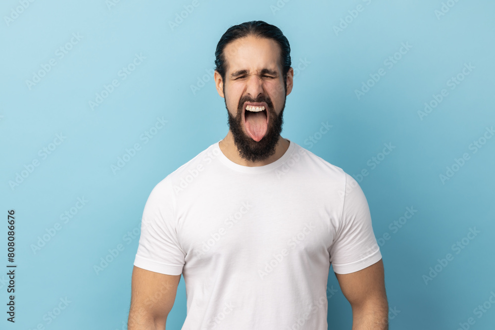 Portrait of playful cheerful man with beard wearing white T-shirt showing tongue, having fun, fooling around, childish manners, keeps eyes closed. Indoor studio shot isolated on blue background. Portrait of playful cheerful man with beard wearing white T-shirt showing tongue, having fun, fooling around, childish manners, keeps eyes closed. Indoor studio shot isolated on blue background.