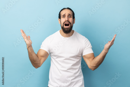 What do you want? Portrait of angry confused man standing with raised hands and surprised indignant expression, asking why how, what reason. Indoor studio shot isolated on blue background.