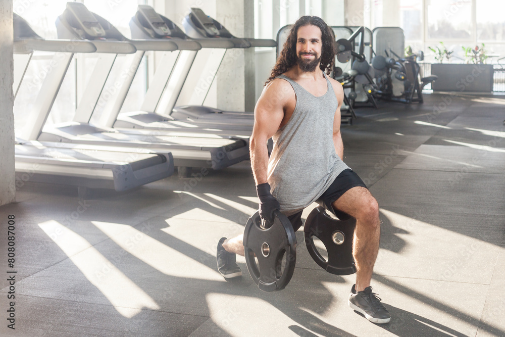 Full length portrait of strong muscular man bodybuilder with long curly hair having workout in gym doing exercises for legs, lunges, holding heavy discs in hands. Indoor shot. Full length portrait of strong muscular man bodybuilder with long curly hair having workout in gym doing exercises for legs, lunges, holding heavy discs in hands. Indoor shot.