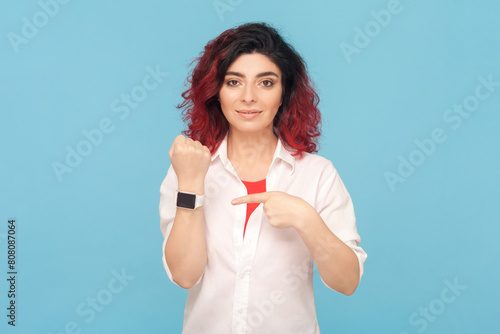Portrait of serious attractive woman with fancy red hair standing pointing at wrist watch, reminding about deadline, wearing white shirt. Indoor studio shot isolated on blue background.