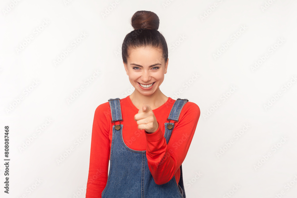 Portrait of joyous pretty attractive woman with hair bun pointing to camera, expresses choice, smiles broadly, wearing denim overalls. Indoor studio shot isolated on white background Portrait of joyous pretty attractive woman with hair bun pointing to camera, expresses choice, smiles broadly, wearing denim overalls. Indoor studio shot isolated on white background