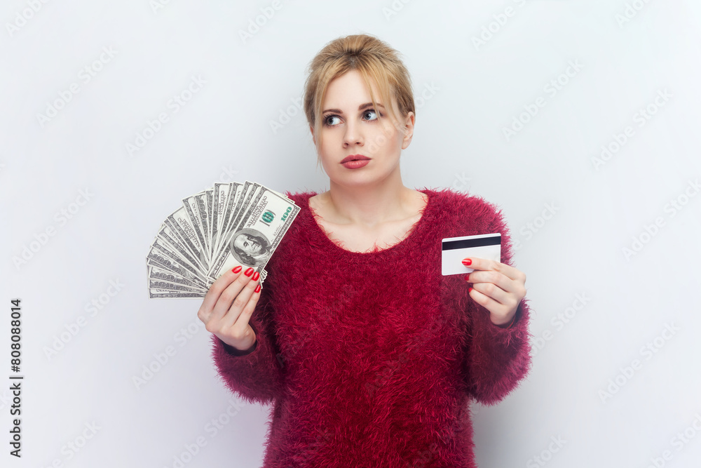 Φωτογραφία Portrait of confused young blonde woman standing holding dollar banknotes and credit card, banking payments, wearing red fluffy jumper