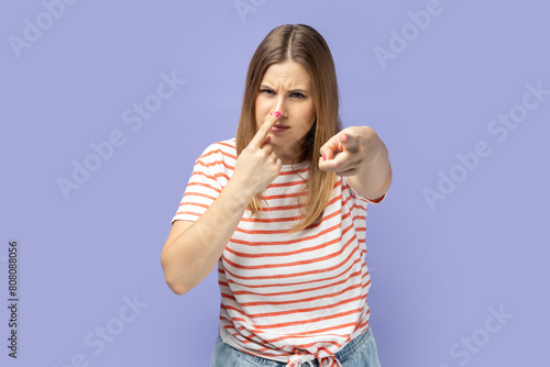 Suspicious blond woman in T-shirt pointing to camera, touching nose, doing liar gesture, body language symbol of cheats, falsehood and deception. Indoor studio shot isolated on purple background. photo