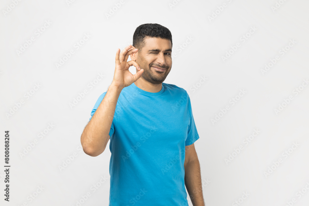 Φωτογραφία Portrait of playful positive optimistic unshaven man wearing blue T- shirt standing looking at camera, showing okay gesture, saying good job