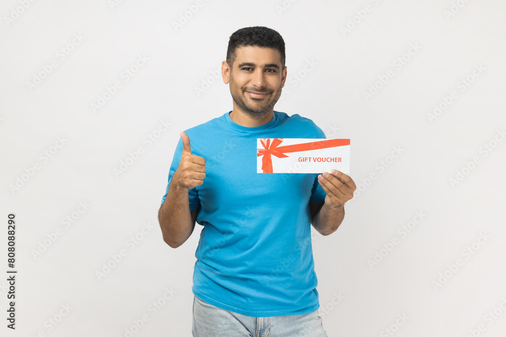 Portrait of handsome delighted satisfied man wearing T- shirt standing with gift voucher and showing thumb up, likes certificate coupon. Indoor studio shot isolated on gray background. Portrait of handsome delighted satisfied man wearing T- shirt standing with gift voucher and showing thumb up, likes certificate coupon. Indoor studio shot isolated on gray background.