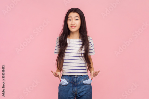 Portrait of poor displeased woman with long brunette hair showing her empty pockets expressing negative emotions, wearing striped T-shirt. Indoor studio shot isolated on pink background. photo