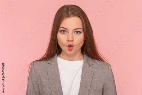 Portrait of childish playful woman with brown hair making fish lips, looking at camera, having fun, grimacing, wearing business suit. Indoor studio shot isolated on pink background. photo