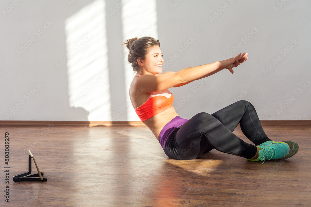 Portrait of athletic woman doing workout watching online lesson with tablet, doing pilates, wearing black sports top and tights. Full length studio shot illuminated by sunlight from window.