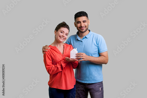 Portrait of loving family man and woman standing together hugging showing paper houise dreaming about home looking at camera. Indoor studio shot isolated on gray background.