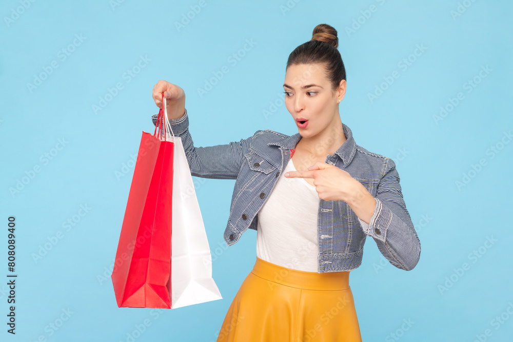 Portrait of surprised shocked woman with bun hairstyle wearing denim jacket pointing at shopping bags amazed by purchases. Indoor studio shot isolated on light blue background. Portrait of surprised shocked woman with bun hairstyle wearing denim jacket pointing at shopping bags amazed by purchases. Indoor studio shot isolated on light blue background.