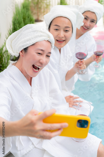 chilling sunday pool party asian adult friend woman in bathrope casual cloth hanging around sit near water pool hand hold glass wine enjoy selfie video calling old friend meeting laugh smile summer