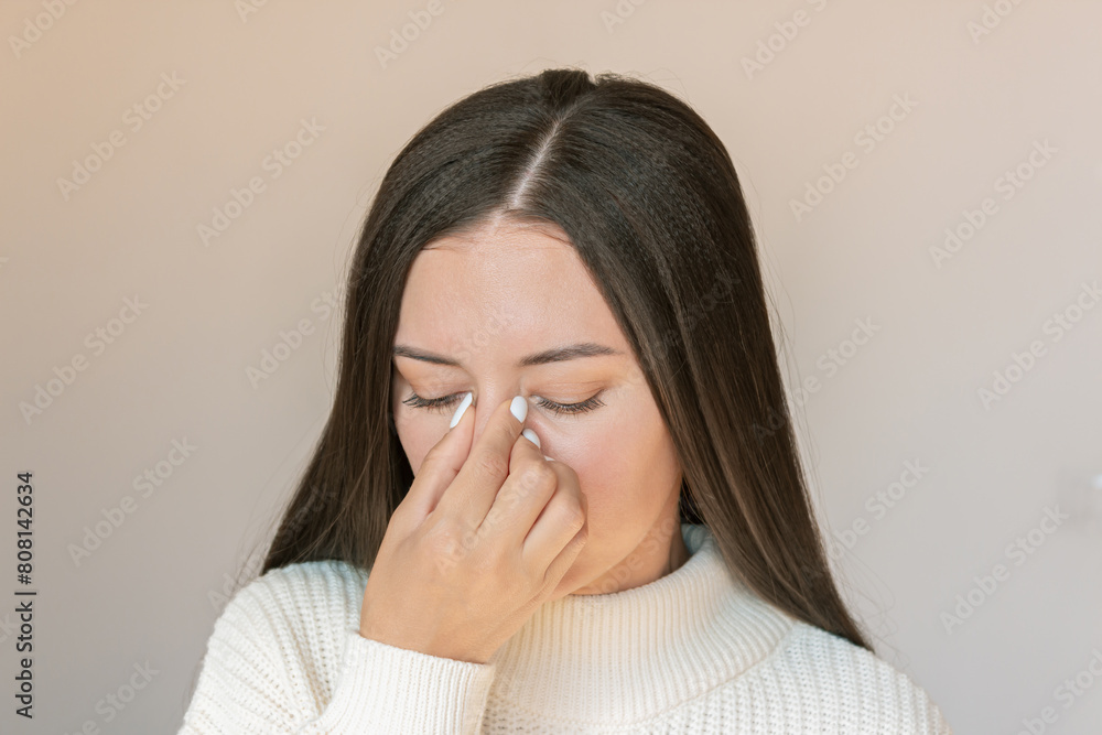 Young Caucasian woman suffers from nasal congestion and runny nose, experiences pain in the nose with sinusitis, holds her nose with her hand. Isolated on a beige background. Allergy Young Caucasian woman suffers from nasal congestion and runny nose, experiences pain in the nose with sinusitis, holds her nose with her hand. Isolated on a beige background. Allergy