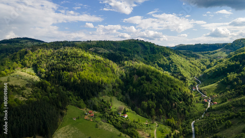 Aerial view of road in green meadows at spring sunny day. Top view from drone of rural road, mountains, forest. Beautiful landscape with roadway, sun rays, trees, hills, green grass, clouds. Serbia © Nenad