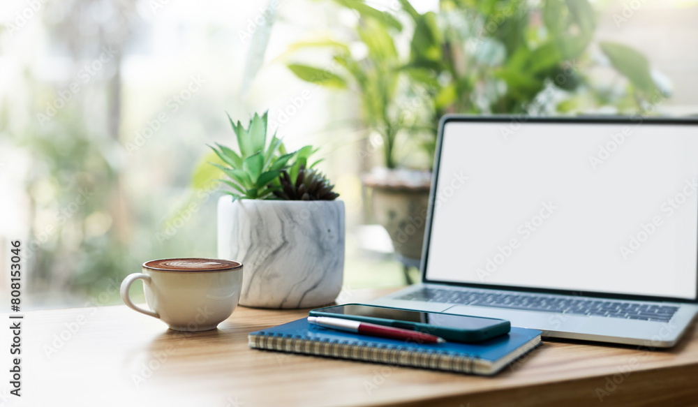 Working space on wooden table. coffee cup with laptop, mobile on notebook and small tree in vase with garden background Working space on wooden table. coffee cup with laptop, mobile on notebook and small tree in vase with garden background