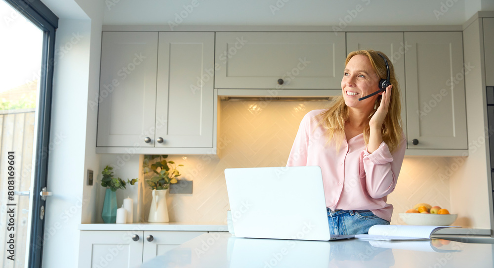Woman Wearing Headset Working From Home In Kitchen Using Laptop Woman Wearing Headset Working From Home In Kitchen Using Laptop