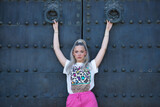 Young, pretty, blonde woman with blue eyes, printed T-shirt and pink skirt, looking at the camera, clinging to some huge knockers of a beautiful old door. Concept of beauty, fashion, millennial, trend