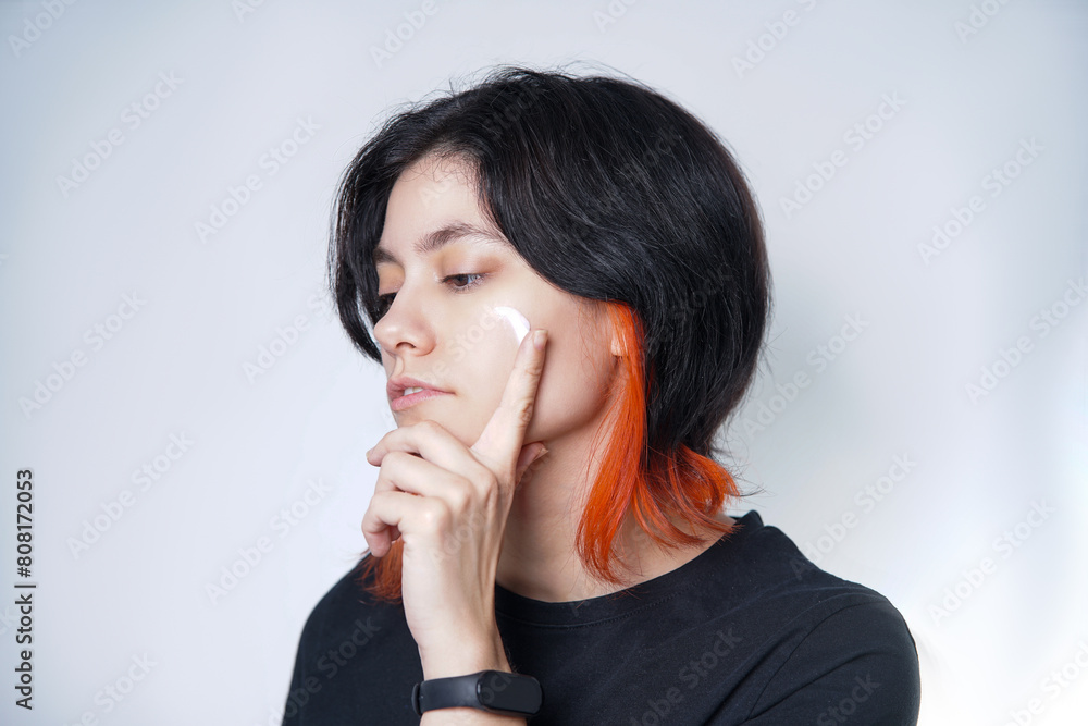 Mediterranean black and orange haired person applying a scoop of moisturizing cream on their cheek with their index finger. Mediterranean black and orange haired person applying a scoop of moisturizing cream on their cheek with their index finger.