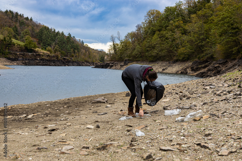 Young female volunteer collecting plastic bottles on a lake shore, with ...