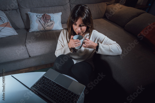 View from above of a young adult woman sitting in the living room with a cup of coffee in her hands, looking at a computer screen under the light of a lamp
