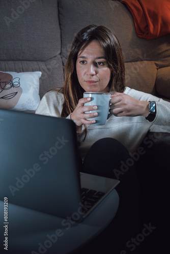 Young adult woman with a cup of coffee in her hands, relaxing in the living room looking at a computer screen under the light of a lamp
