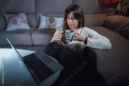 Young adult woman with a cup of coffee in her hands, relaxing in the living room looking at the computer under the light of a lamp
