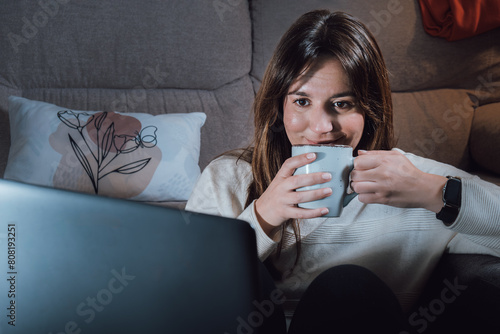 Young adult woman with a cup of coffee in her hands, relaxing in the living room looking at the computer
