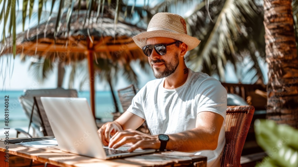 A man in a straw hat sits before a laptop at a table Palm trees frame the background A man in a straw hat sits before a laptop at a table Palm trees frame the background