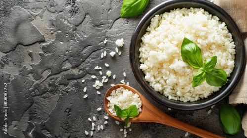  A bowl of rice on a gray surface, garnished with basil leaves A wooden spoon rests nearby, with a cloth within reach