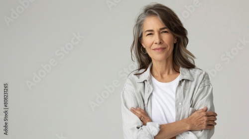 Mature woman with gray hair, arms crossed, wearing a white shirt, confident and serene, on a light background © Iona