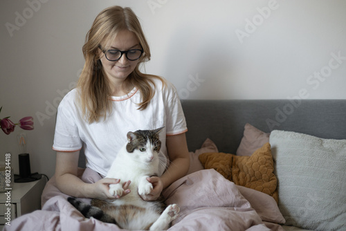Happy young 30s woman with cat in bed at home. In cold weather, Pet friendly concept. Stray kitten sleep on bed. Cozy home background, morning bedtime.