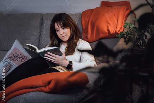Young adult woman reading a book, relaxed on the sofa in her living room under the light of the lamp