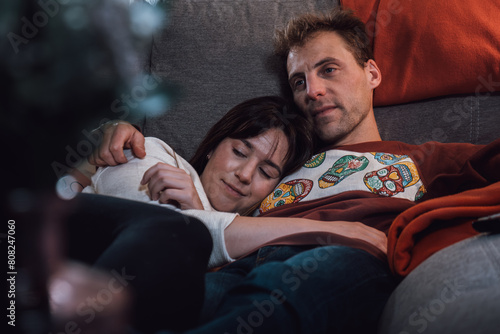 Couple of young adult man and woman lying and hugging while resting on the couch. Real life people at home
