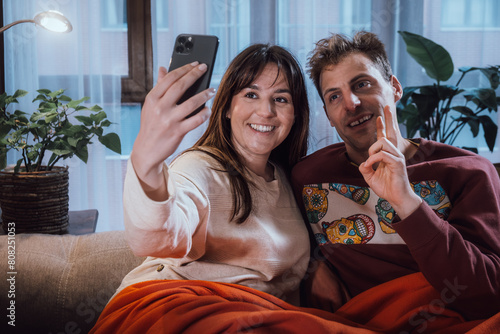 Young adult man and woman couple making a video call or posing for a selfie on the couch in their living room. Real life people at home
