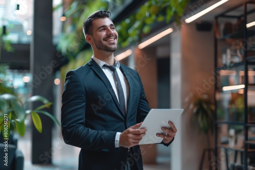 Young business man CEO wearing suit standing in office using digital tablet. Smiling mature businessman professional executive manager looking away thinking working on tech device